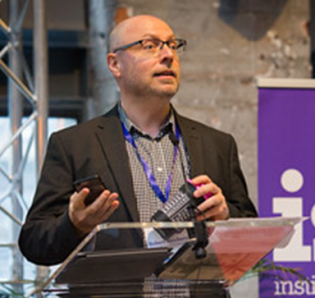 A photo of Dr Robert Newbery speaking behind a podium at an event. He is the Keynote Speaker at REC2023, and Professor of Entrepreneurship at Northumbria University.