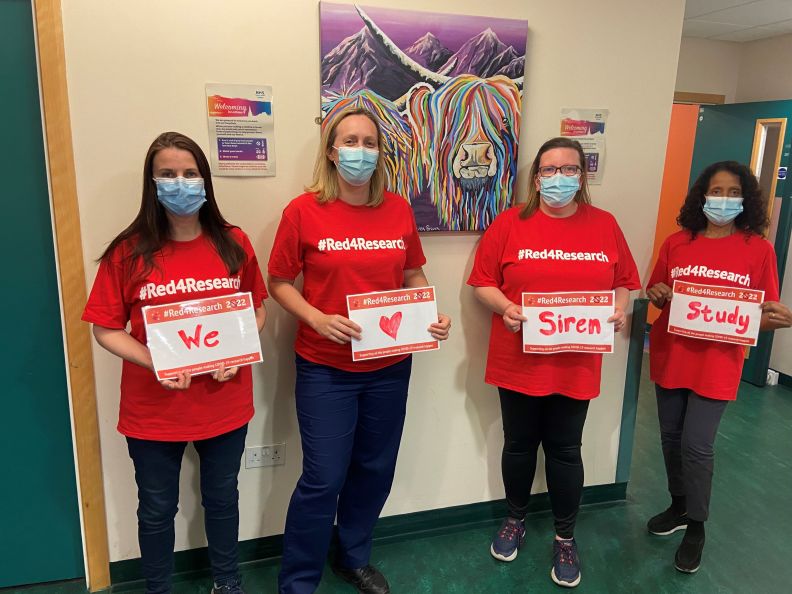 Four people standing in a corridor in front of a wall wearing NHS Lothian red for research t-shirts, holding signs that read we heart siren study.