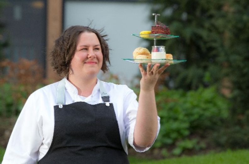 A profile image of Helen Vass, a GCU alumna and a pastry chef, in chef's uniform holding a selection of cakes in a cake stand.