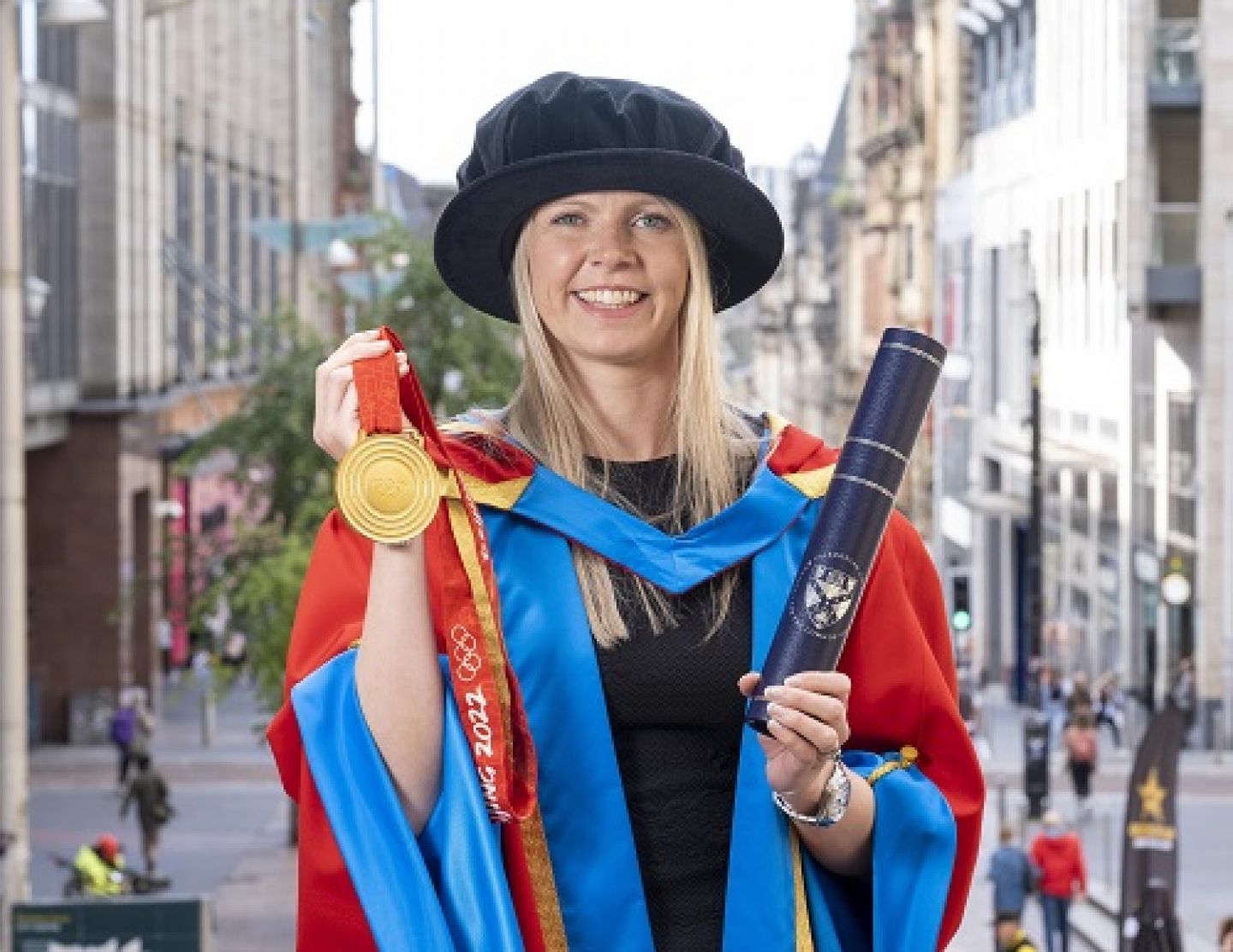 A profile picture of Vicky Wright, a GCU alumna, Honorary Graduate, and a Scottish curler, in her graduation robes and cap, holding her Olympic Gold medal and her Honorary Degree container.