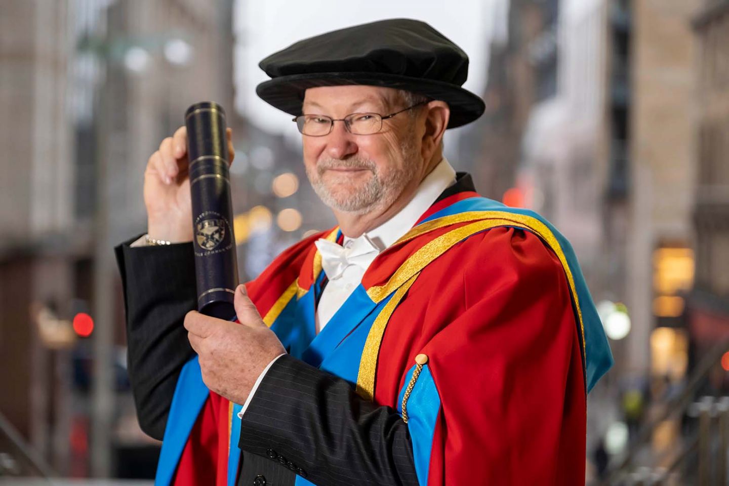 GCU Honorary Graduate Dr Jim Paterson wearing his graduation hood and hat and holding a degree container