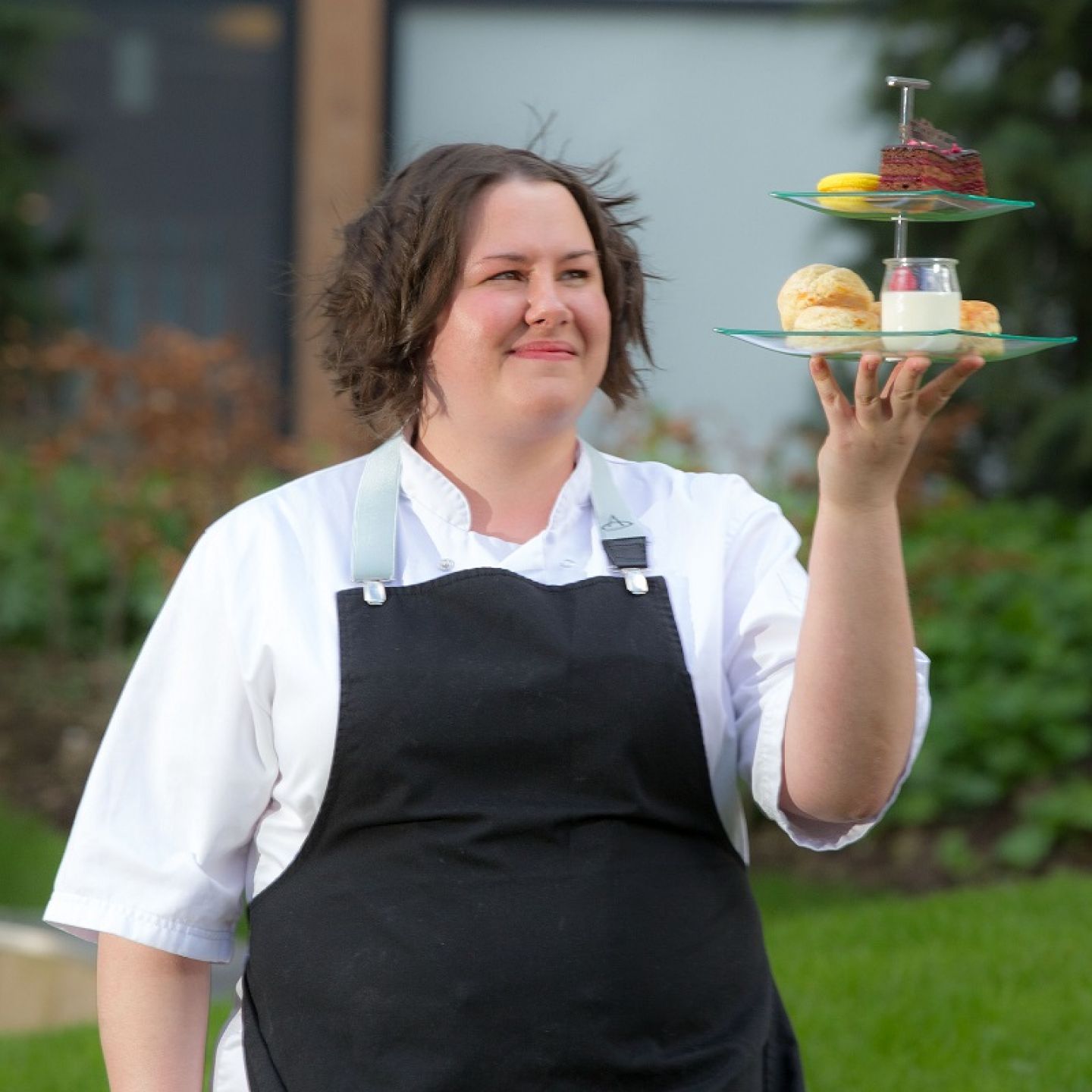 A profile picture of Helen Vass, a GCU alumna and a pastry chef, in chef's uniform holding a selection of cakes in a cake stand.