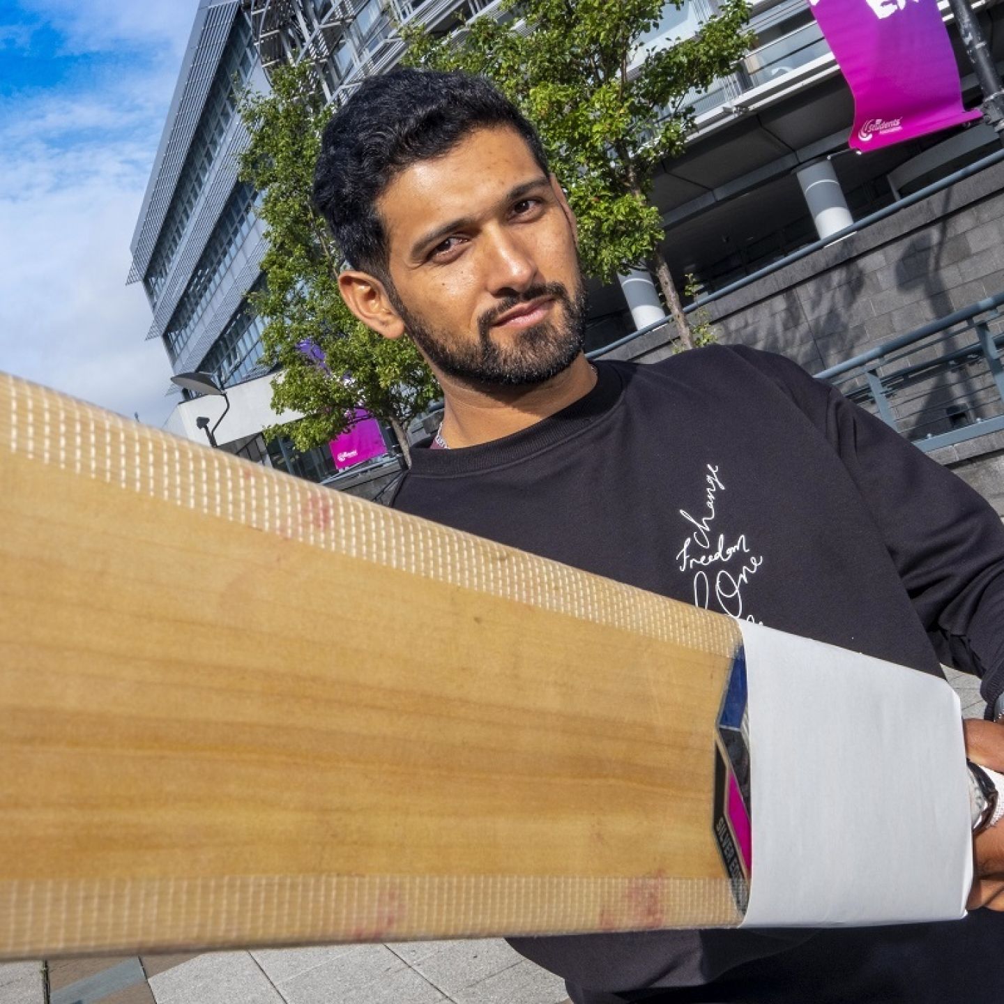 A profile picture of Sikandar Raza, a GCU alumnus and an international cricketer, holding a cricket bat while standing in the exterior of Glasgow campus.