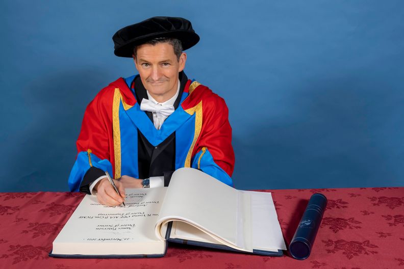 GCU Honorary Graduate Dr Anthony Burns signing a book in gradauation hood and hat.
