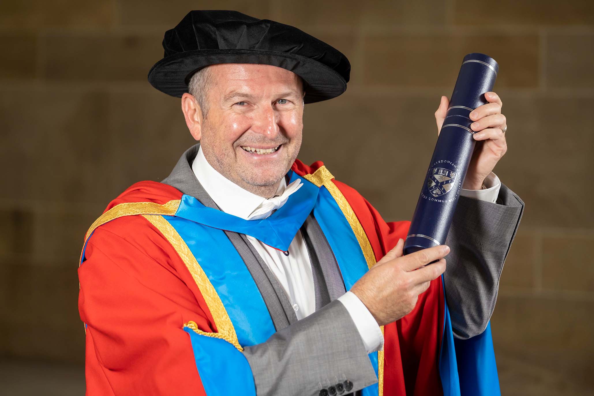 GCU Honorary Graduate Dr Stephen Breslin wearing his graduation hood and hat and holding a degree container.