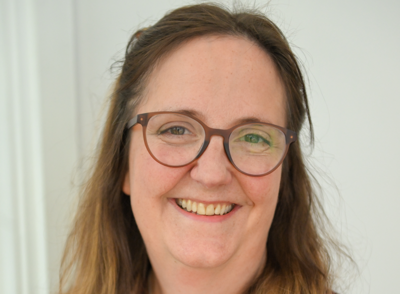 A white female with shoulder-length brown hair and brown glasses smiles at the camera.
