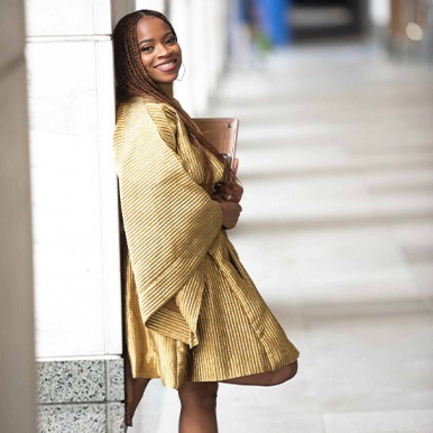 A square photo of Abi Daré, GCU alumna and New York Times bestselling author standing against a column, holding a laptop and smiling.