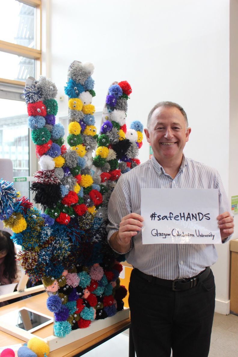 Person standing in front of large sculpture of hand covered in multi-coloured germs, holding a sign that reads #safeHANDS Glasgow Caledonian University